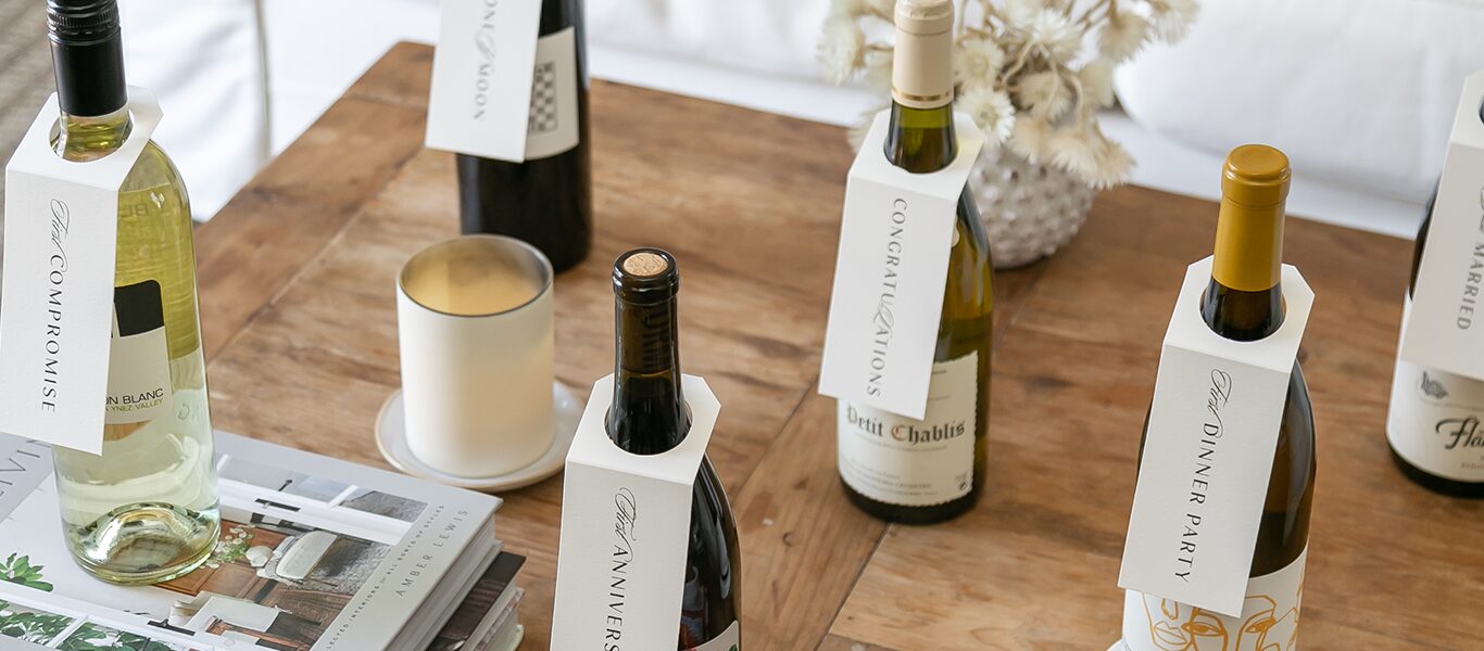 Wine gift bottles with celebration occasion tags on a wooden table with a candle and decorative elements.