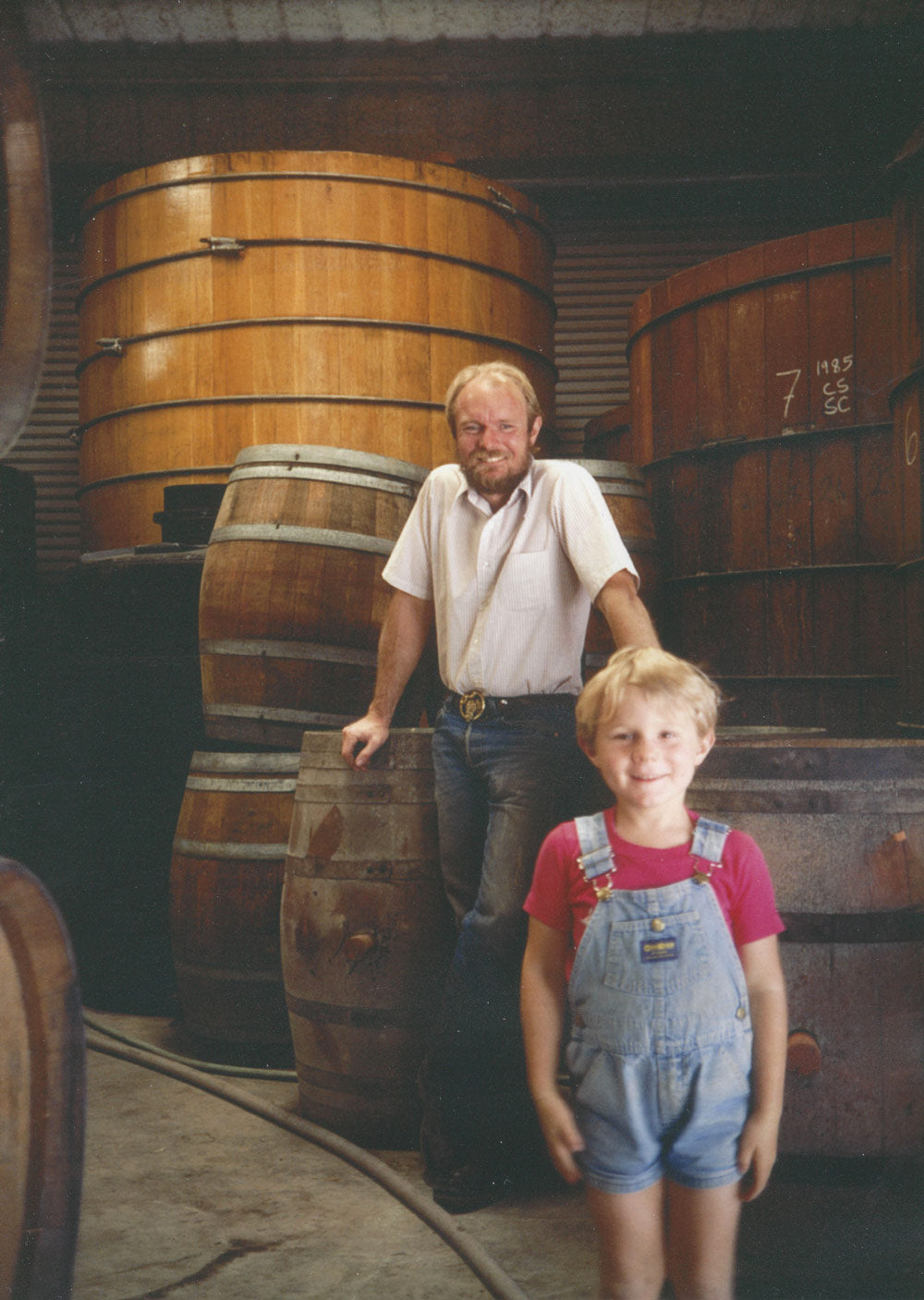 Vintage photo of a winemaker and a young boy in overalls standing among large oak barrels in a cellar