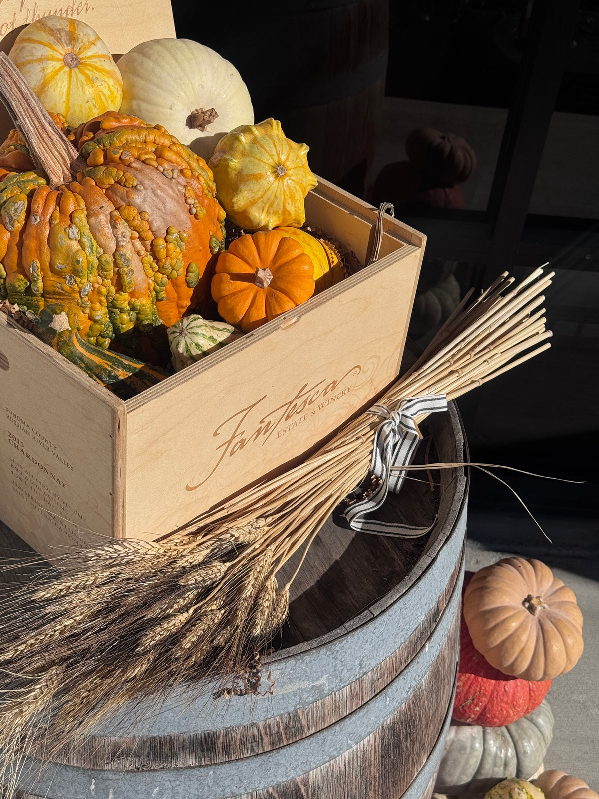 Wooden Fantesca wine crate filled with colorful fall gourds and pumpkins beside a bundle of wheat on a barrel
