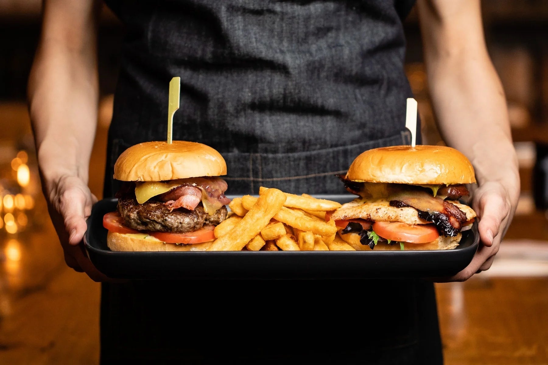 Person holding a tray with two burgers and fries in an indoor setting