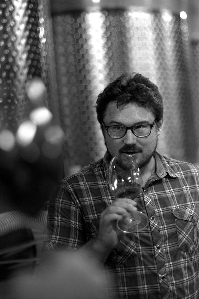 Black-and-white portrait of a winemaker holding a glass of red wine in front of a steel fermentation tank